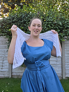 Woman in a blue dress smiles while holding up white lace knitted scarf with pink ribbons woven through in a leafy garden.