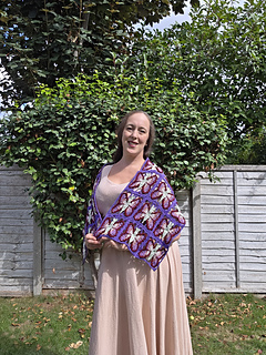 crochet lace shawl with colourful butterfly design  in blues purples, pinks and green on a woman in a leafy garden (close, front-side)