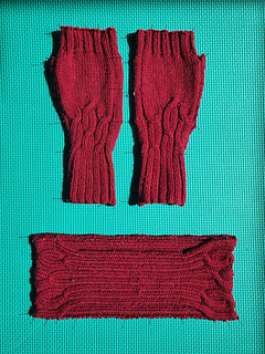 Deep red cable knitted headband and matching pair of fingerless gloves (not included in Headband pattern) pinned on a green foam mat.