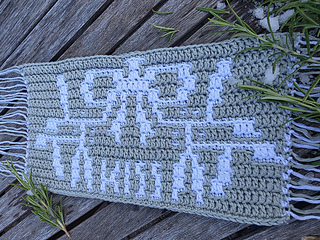 Crocheted green and white mosaic panel with spray of mistletoe tied with a bow and text spelling KISS in white on a weathered wooden deck, with rosemary sprigs and melting snow.