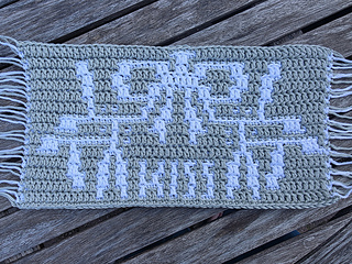 Crocheted green and white mosaic panel with spray of mistletoe tied with a bow and text spelling KISS in white on a weathered wooden deck, with rosemary sprigs and melting snow.