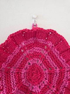 Red knitted circular washcloth with pink stripes with flowers hanging on a hook on a white wall.