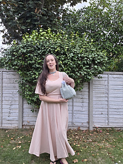 Smiling woman outdoors holds a pale sage green crocheted tote with mistletoe design and KISS text in white, standing before a leafy fence and trees.