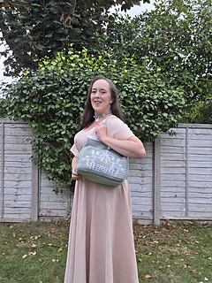 Smiling woman outdoors holds a pale sage green crocheted tote with mistletoe design and KISS text in white, standing before a leafy fence and trees.