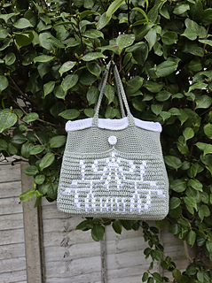 A pale sage green crocheted tote with mistletoe design and KISS text in white, hung on a leafy hedge.