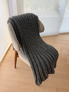 The Cloud Blanket — charcoal wool, draped over a woven chair.