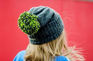 View of a blonde woman's back of head wearing a blue slouch hat with ribbed brim folded up and a bright green pom pom.