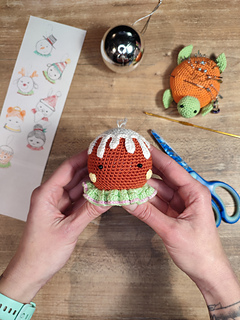 Crocheted gingerbread brown head, white icing details, and colorful decorative buttons. Held in the hands, crochet tools put on the table, scissors, crochet hook, plain Christmas bauble.
