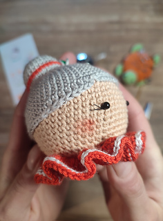 Crochet Mrs. Claus doll wearing a red collar with white details and gray hair in a bun. Face detail, right head side.