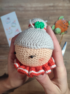 Crochet Mrs. Claus doll wearing a red collar with white details and gray hair in a bun. Face detail, upper head, left head side.