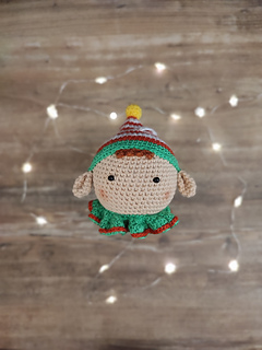 Handmade crochet Christmas elf with pointed hat, striped outfit, and playful expression. Christmas lights visible in the background.