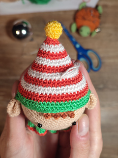 Handmade crochet Christmas elf with pointed hat, striped outfit, and playful expression. Held in the hands. Crochet tools visible in the background. Detailed hat and top of the head view.