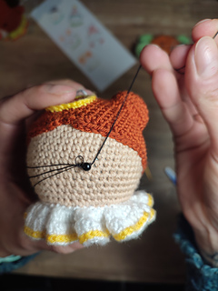 Crochet amigurumi style Christmas bauble. Brown hair with two buns, yellow halo at the top of the head and white-yellow collar. Embroidering the eyelashes onto the angel's face.