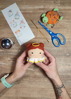 Crochet amigurumi style Christmas bauble. Brown hair with two buns, yellow halo at the top of the head and white-yellow collar. Held in the hands. Crafting tools are on the table in the background, Christmas bauble, scissors, hook, pins and hand drawn sketches.
