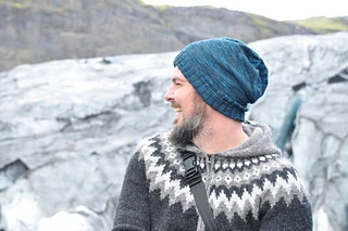 Man with beard laughing and wearing a blue slouchy knit hat and a fairisle sweater in grey tones standing in front of rocks.