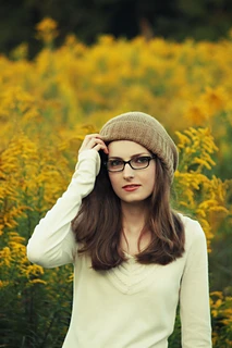 Brunette woman with long hair and beige long-sleeved shirt wearing glasses and a fawn-coloured slouchy knit hat with the ribbed brim folded up and goldenrod in the background.