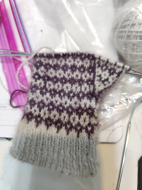 The beginnings of a mitten with white 1x1 ribbing and a colorwork pattern in purple. The pattern is a bunch of 6 row diamonds? I think? I don't know how to describe it. The thumb gusset has reversed color dominance and is outlined in a line of purple and a line of white. It's all on two flexiflip needles.