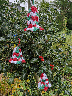 Red, green and white crocheted Christmas bell ornaments hanging in a holly bush with red berries