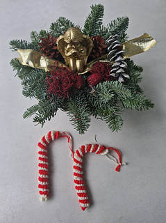 Crocheted red and white candy cane ornaments lying next to a Christmas fir arrangement with pine cones and gold decoration